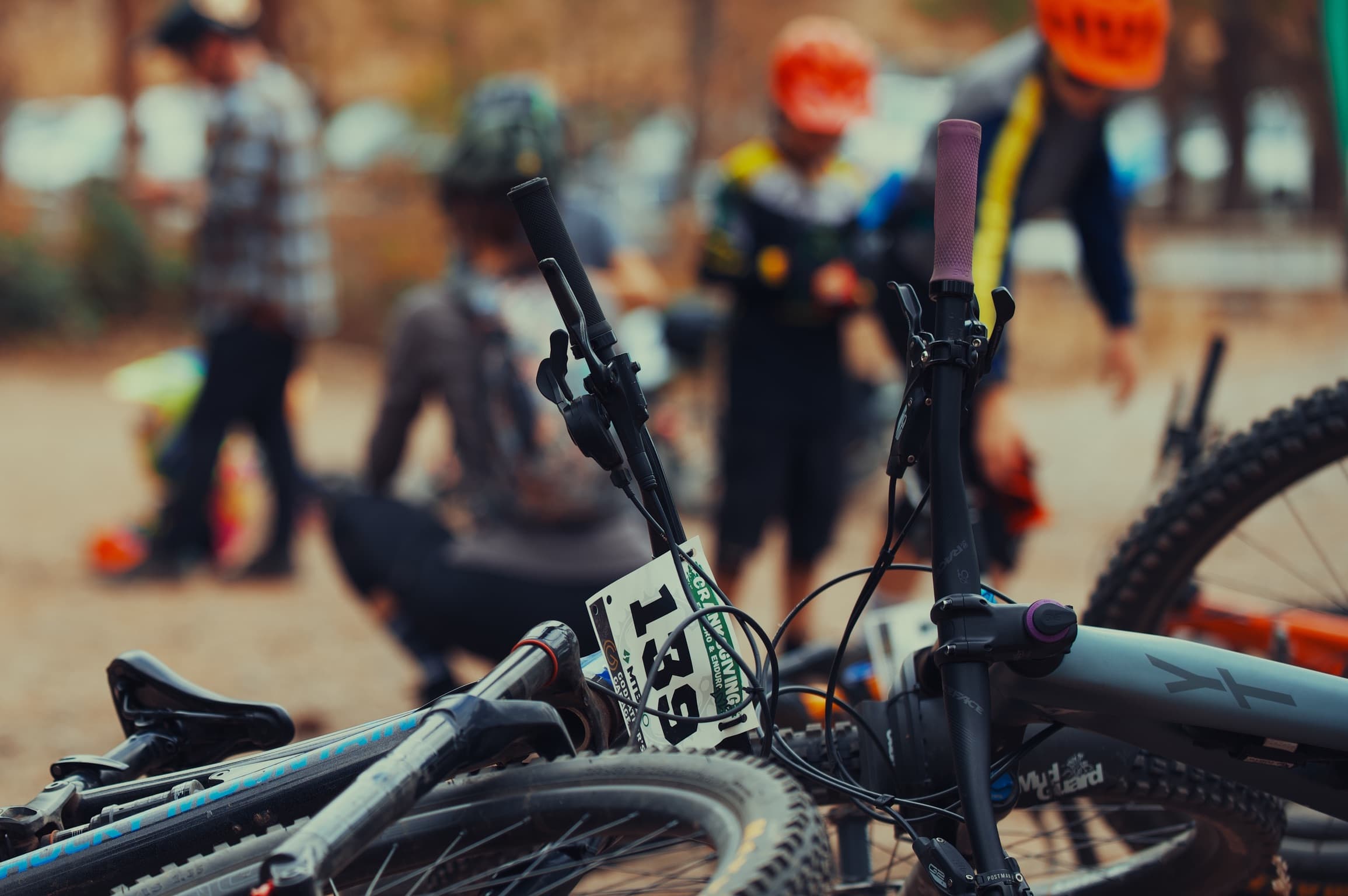 Mountain bikes laid on the ground with race plates visible, riders in helmets in the background