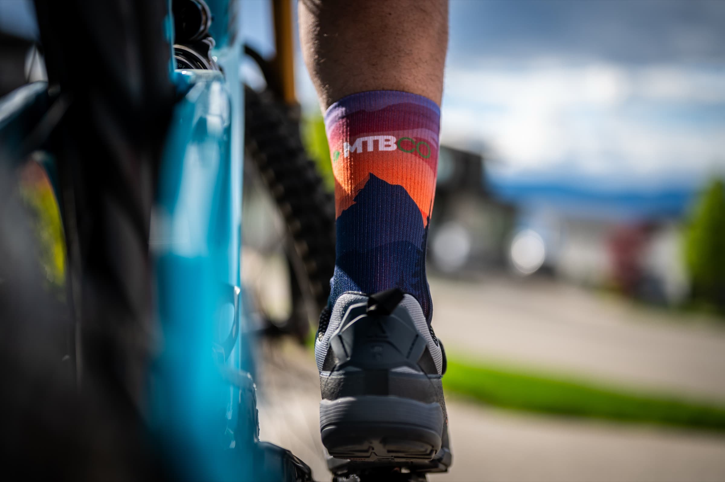 Cyclist's foot on a pedal wearing MTBCO socks with sunset mountain graphic, lake visible in the background